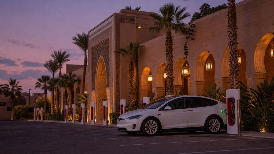 Белая Tesla Model X заряжается у суперчарджера на пальмовой парковке марокканского отеля в сумерках